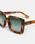 Nahaufnahme einer quadratischen Sonnenbrille im Schildpatt-Look mit dunklen, verlaufenden, polarisierten Gläsern. Die Sonnenbrille verfügt über einen breiten, braun-bernsteinfarben marmorierten Rahmen im Oversize-Design; auf dem linken Bügel ist „Okkia“ in Weiß aufgedruckt. Der Hintergrund ist schlicht weiß, wodurch diese stilvolle Okkia Club Sonnenbrille besonders gut zur Geltung kommt.