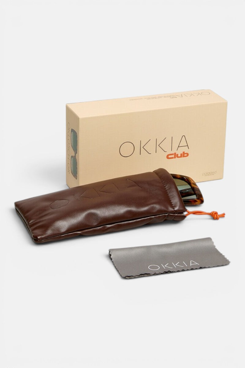 Auf einer weißen Fläche liegen eine beigefarbene Okkia Club-Box, ein braunes, gepolstertes Sonnenbrillenetui mit orangefarbenem Kordelzug, ein graues Okkia-Reinigungstuch sowie die Oversize-Sonnenbrille „ISCHIA“ mit polarisierten Gläsern und sichtbaren Bügeln.