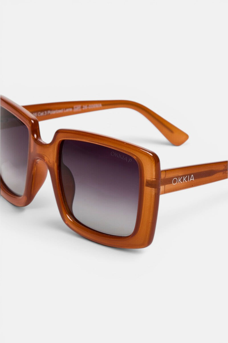Nahaufnahme der Okkia ISCHIA Sonnenbrille mit einem markanten, quadratischen Rahmen in kräftigem, durchscheinendem Orange und dunklen Verlaufsglasgläsern. Das Modell im Oversize-Design trägt den weißen Okkia-Schriftzug auf dem Bügel. Die Sonnenbrille wird vor einem schlichten weißen Hintergrund präsentiert, um die Details besonders hervorzuheben.