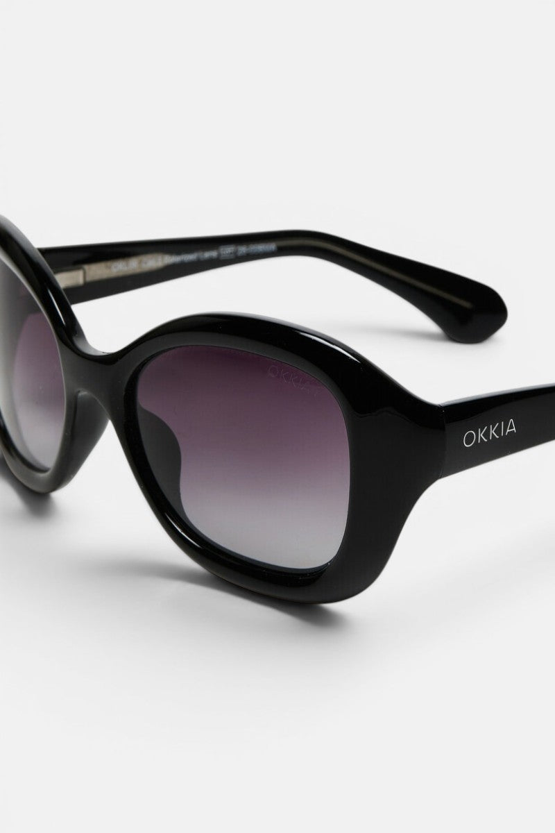 Schwarze, ovale Retro-Sonnenbrille mit dunklen Verlaufsgläsern und einem markanten, breiten Rahmen. Der Markenname „Okkia“ ist in Weiß auf der Außenseite des linken Bügels aufgedruckt und verweist damit auf die Verbindung zum Okkia Club. Diese polarisierte Sonnenbrille wird vor einem schlichten, hellgrauen Hintergrund präsentiert.