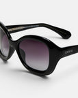 Schwarze, ovale Retro-Sonnenbrille mit dunklen Verlaufsgläsern und einem markanten, breiten Rahmen. Der Markenname „Okkia“ ist in Weiß auf der Außenseite des linken Bügels aufgedruckt und verweist damit auf die Verbindung zum Okkia Club. Diese polarisierte Sonnenbrille wird vor einem schlichten, hellgrauen Hintergrund präsentiert.