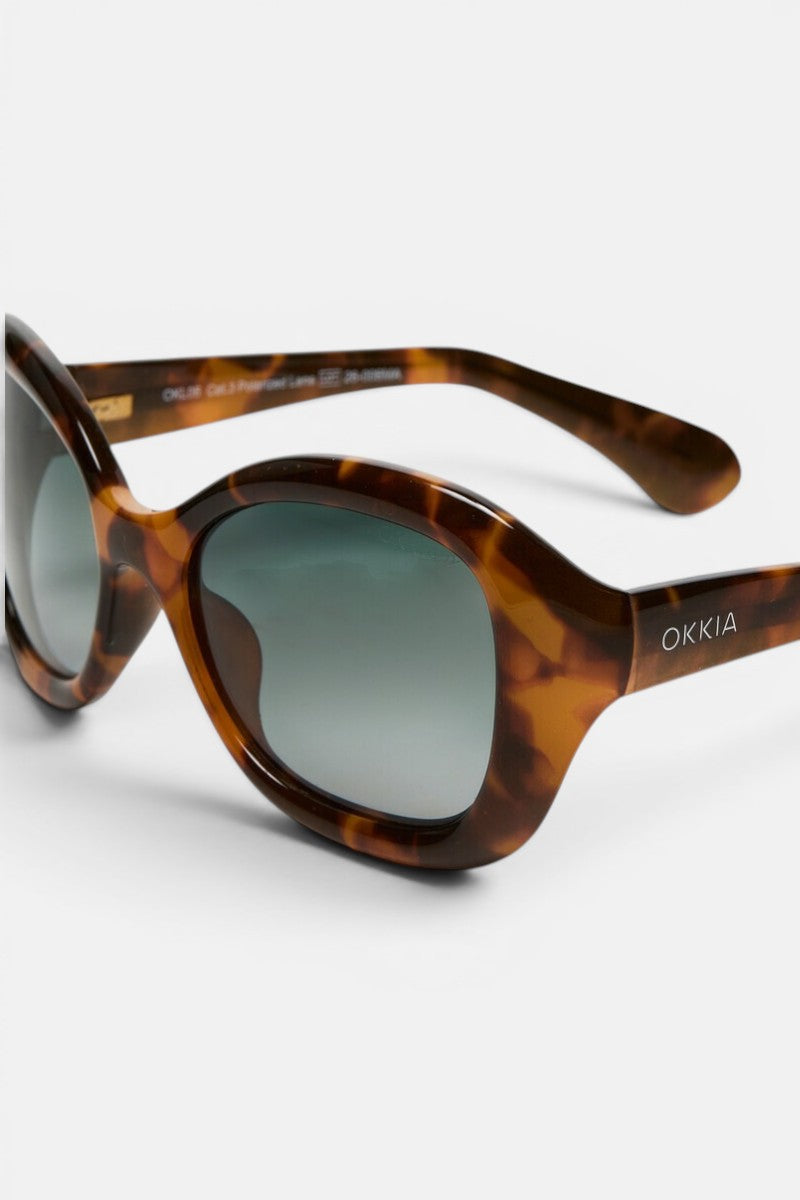 Diese braune Sonnenbrille im Schildpatt-Look mit ovalen Gläsern und hellblauem Farbverlauf verkörpert den Retro-Stil von Okkia. Das Gestell ist am Bügel mit einer weißen „OKKIA“-Schrift verziert, die den charakteristischen Stil der Marke widerspiegelt. Diese polarisierte Sonnenbrille liegt auf einer weißen Unterlage und ist so positioniert, dass sowohl die Vorderseite als auch das Seitenprofil gut zur Geltung kommen.