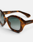 Diese braune Sonnenbrille im Schildpatt-Look mit ovalen Gläsern und hellblauem Farbverlauf verkörpert den Retro-Stil von Okkia. Das Gestell ist am Bügel mit einer weißen „OKKIA“-Schrift verziert, die den charakteristischen Stil der Marke widerspiegelt. Diese polarisierte Sonnenbrille liegt auf einer weißen Unterlage und ist so positioniert, dass sowohl die Vorderseite als auch das Seitenprofil gut zur Geltung kommen.