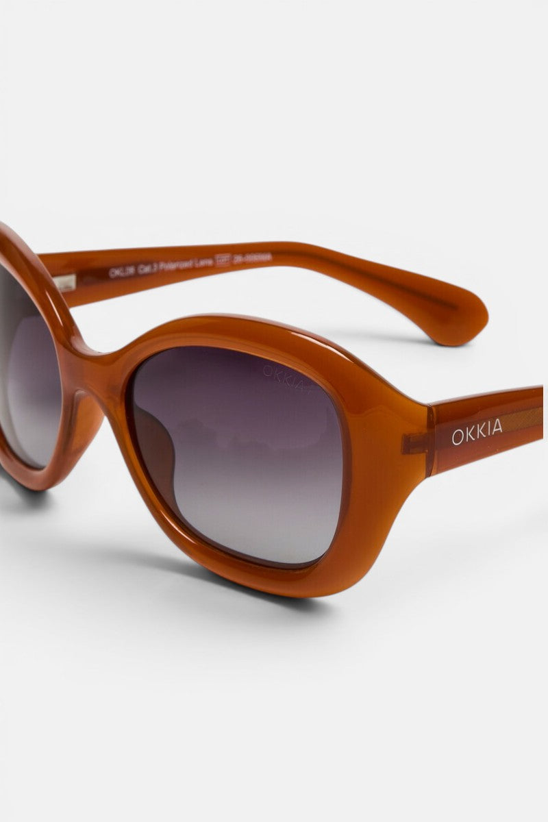 Nahaufnahme einer braunen, ovalen Okkia-Sonnenbrille im Retro-Stil mit breitem Rahmen und dunkel verlaufenden Gläsern. Auf der Innenseite des Bügels sind Produktdetails zu sehen, während der äußere Bügel mit dem Schriftzug „Okkia“ in Weiß verziert ist, der den unverwechselbaren Stil von Okkia CLUB widerspiegelt. Diese polarisierte Sonnenbrille hebt sich deutlich von einem schlichten weißen Hintergrund ab.