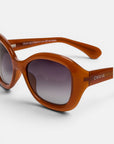 Nahaufnahme einer braunen, ovalen Okkia-Sonnenbrille im Retro-Stil mit breitem Rahmen und dunkel verlaufenden Gläsern. Auf der Innenseite des Bügels sind Produktdetails zu sehen, während der äußere Bügel mit dem Schriftzug „Okkia“ in Weiß verziert ist, der den unverwechselbaren Stil von Okkia CLUB widerspiegelt. Diese polarisierte Sonnenbrille hebt sich deutlich von einem schlichten weißen Hintergrund ab.