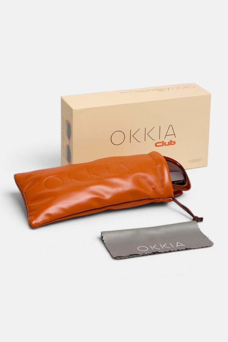 Auf einem weißen Hintergrund sind eine hellbraune Schachtel mit der Aufschrift „Okkia Club“, ein braunes Sonnenbrillenetui mit dem geprägten Schriftzug „Okkia“, ein graues Putztuch mit dem Schriftzug „Okkia“ sowie eine polarisierte Sonnenbrille, die teilweise aus dem Etui herausragt, sind vor einem weißen Hintergrund angeordnet – perfekt für Fans von Okkia und Retro-Brillen.