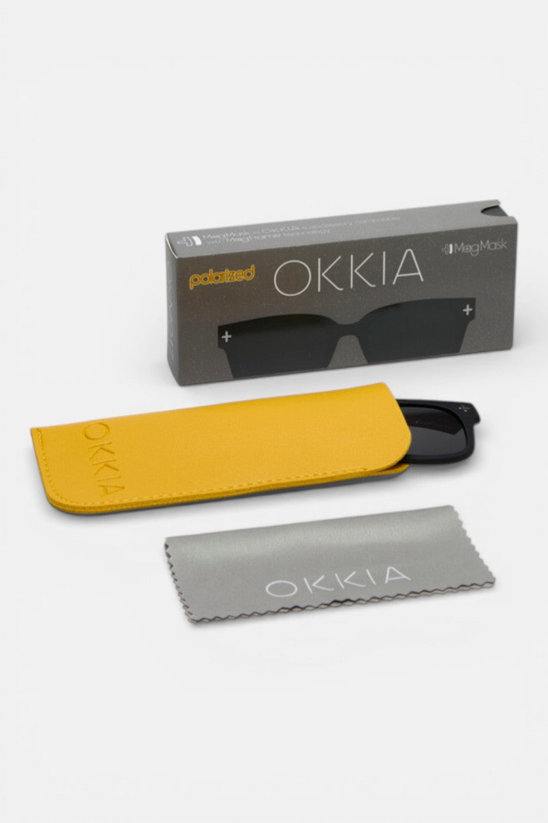 Eine schwarze polarisierte Okkia-Sonnenbrille, teilweise in einem gelben Etui, mit einem grauen Brillenglastuch und einer grauen Okkia-Markenschachtel mit der Aufschrift "polarized", ist auf einer weißen Fläche angeordnet.