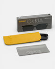 Eine schwarze polarisierte Okkia-Sonnenbrille, teilweise in einem gelben Etui, mit einem grauen Brillenglastuch und einer grauen Okkia-Markenschachtel mit der Aufschrift "polarized", ist auf einer weißen Fläche angeordnet.