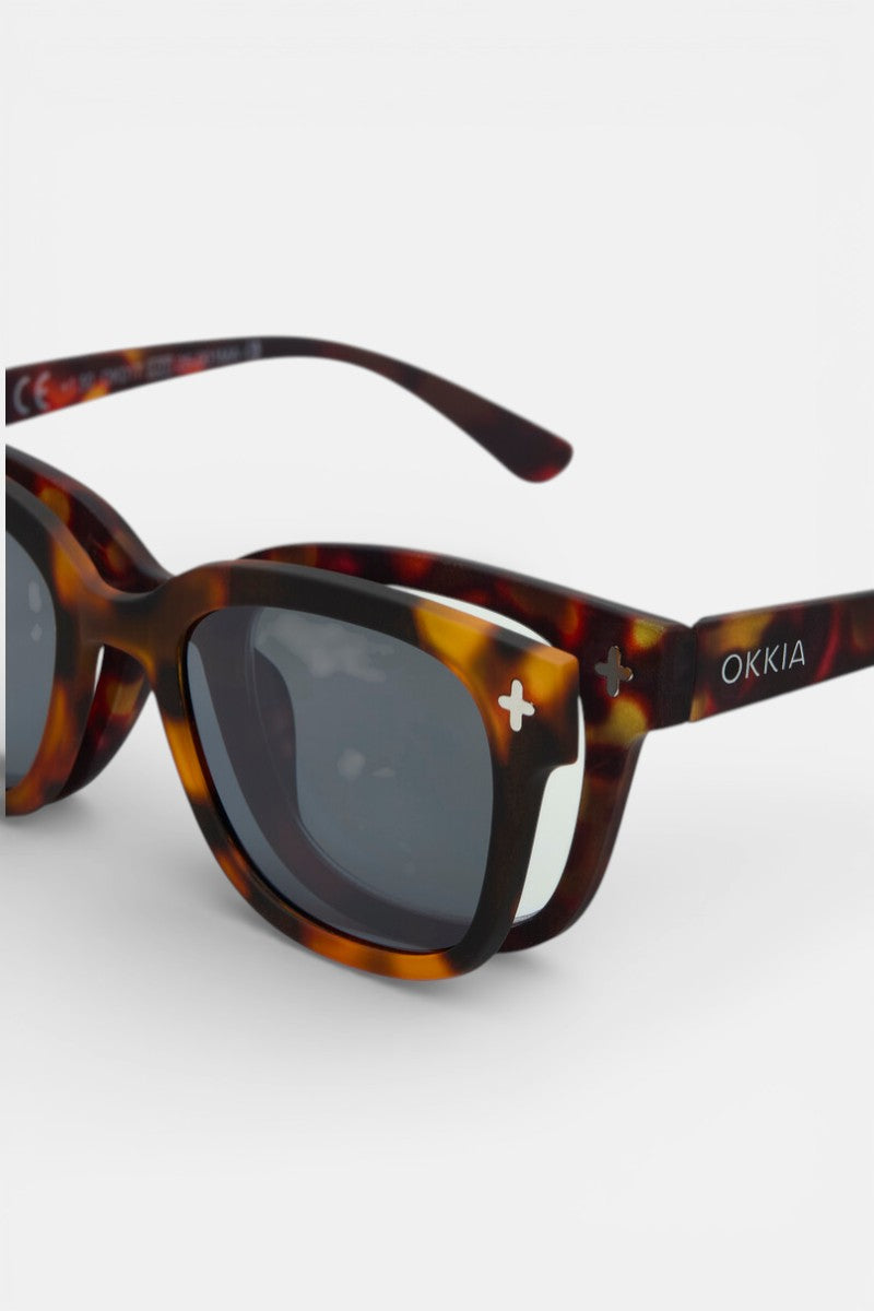 Nahaufnahme einer braunen Schildpatt-Sonnenbrille von Okkia mit rechteckigen dunklen Gläsern, kleinen weißen Pluszeichen an den vorderen Ecken und OKKIA-Branding am Bügel. Der Rahmen ähnelt einer polarisierten Sonnenbrille. Vor einem schlichten weißen Hintergrund.