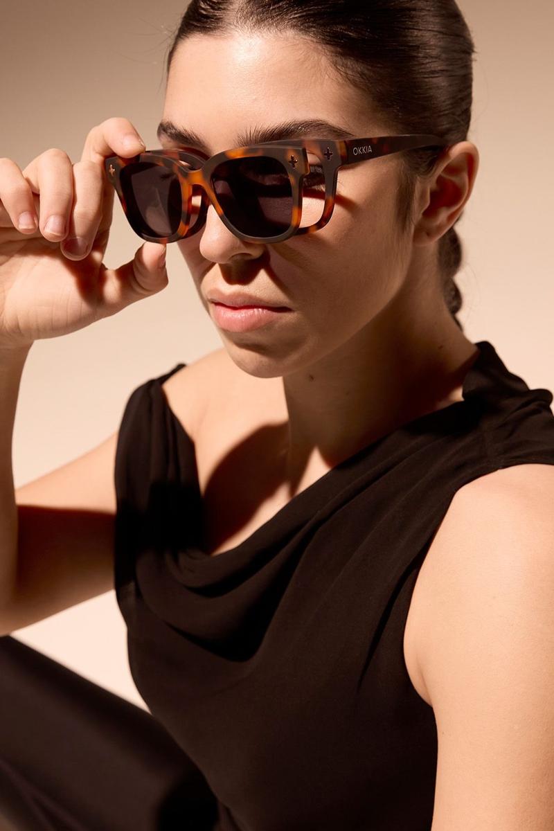 Eine Frau mit dunklem Haar trägt ein ärmelloses schwarzes Oberteil und eine polarisierte Sonnenbrille von Okkia. Sie blickt ernst nach vorne, während sie mit einer Hand die Sonnenbrille vor einem sanft beleuchteten neutralen Hintergrund hält.