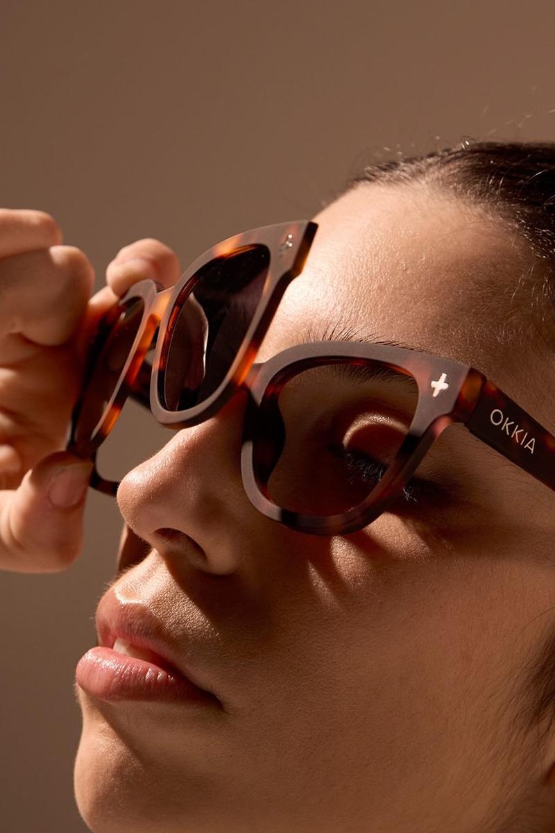 Nahaufnahme einer Person, die eine braune schildpattfarbene Okkia-Sonnenbrille mit der Aufschrift "Okkia" auf dem Bügel trägt. Ihre Hand rückt das Gestell zurecht, und das warme Licht hebt die glatte Haut und den neutralen Gesichtsausdruck hervor.