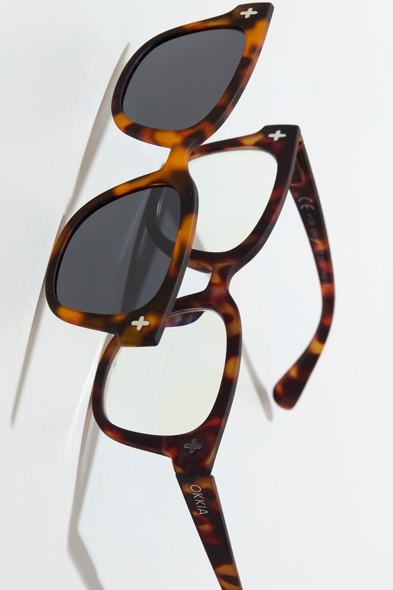 Zwei Paar Okkia Schildpatt-Sonnenbrillen in Katzenaugenform sind übereinander gestapelt; die obere hat polarisierte Gläser, die untere hat klare Gläser. Jede hat ein kleines weißes Kreuz in der Ecke und ein "Okkia"-Branding am Bügel.