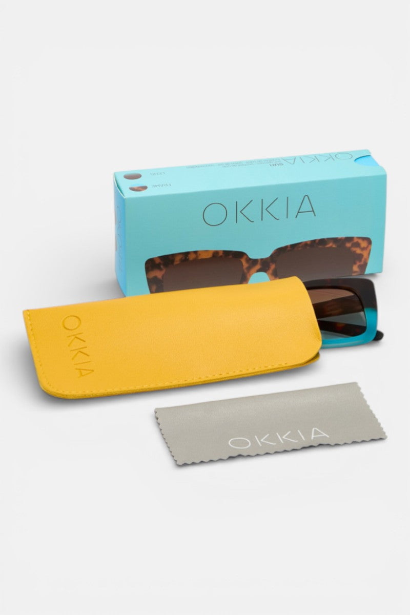 Eine Okkia-Sonnenbrille mit schildpattfarbenem Oversize-Rahmen sitzt teilweise in einem gelben Okkia-Etui. Eine hellblaue Okkia-Box und ein graues "Okkia"-Reinigungstuch sind ebenfalls vor einem weißen Hintergrund abgebildet.