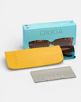 Eine Okkia-Sonnenbrille mit schildpattfarbenem Oversize-Rahmen sitzt teilweise in einem gelben Okkia-Etui. Eine hellblaue Okkia-Box und ein graues "Okkia"-Reinigungstuch sind ebenfalls vor einem weißen Hintergrund abgebildet.
