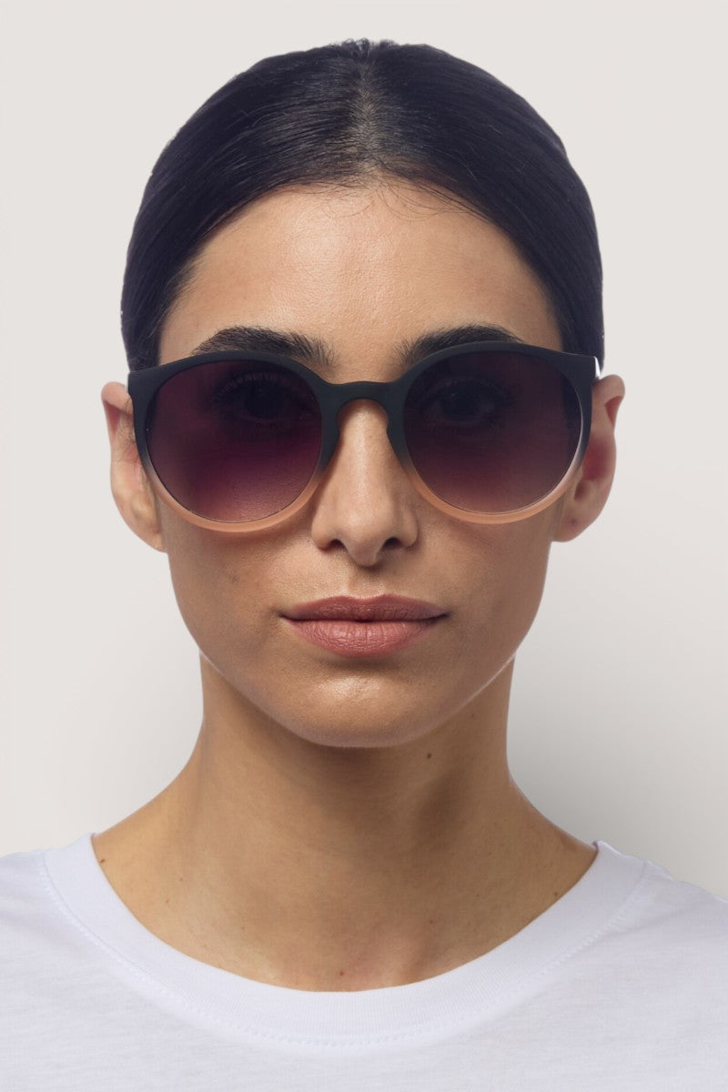 Eine Person mit glattem, dunklem Haar trägt ein weißes Hemd mit Rundhalsausschnitt und eine runde Sonnenbrille von Okkia Berlino mit Verlaufsgläsern und UV-400-Schutz vor einem schlichten, hellen Hintergrund mit neutralem Ausdruck.