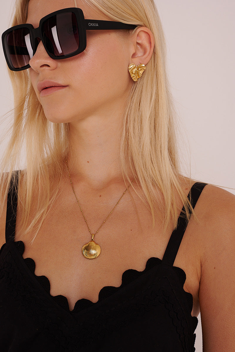 Eine blonde Frau mit einer großen schwarzen Sonnenbrille, einem schwarzen Oberteil mit Wellenschliff und einer goldenen Muschelkette trägt den Princess Goes Hollywood Edelstahl Ohrstecker mit einer organischen Form, ihr glattes Haar liegt offen vor einem schlichten hellen Hintergrund.