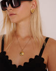 Eine blonde Frau mit einer großen schwarzen Sonnenbrille, einem schwarzen Oberteil mit Wellenschliff und einer goldenen Muschelkette trägt den Princess Goes Hollywood Edelstahl Ohrstecker mit einer organischen Form, ihr glattes Haar liegt offen vor einem schlichten hellen Hintergrund.