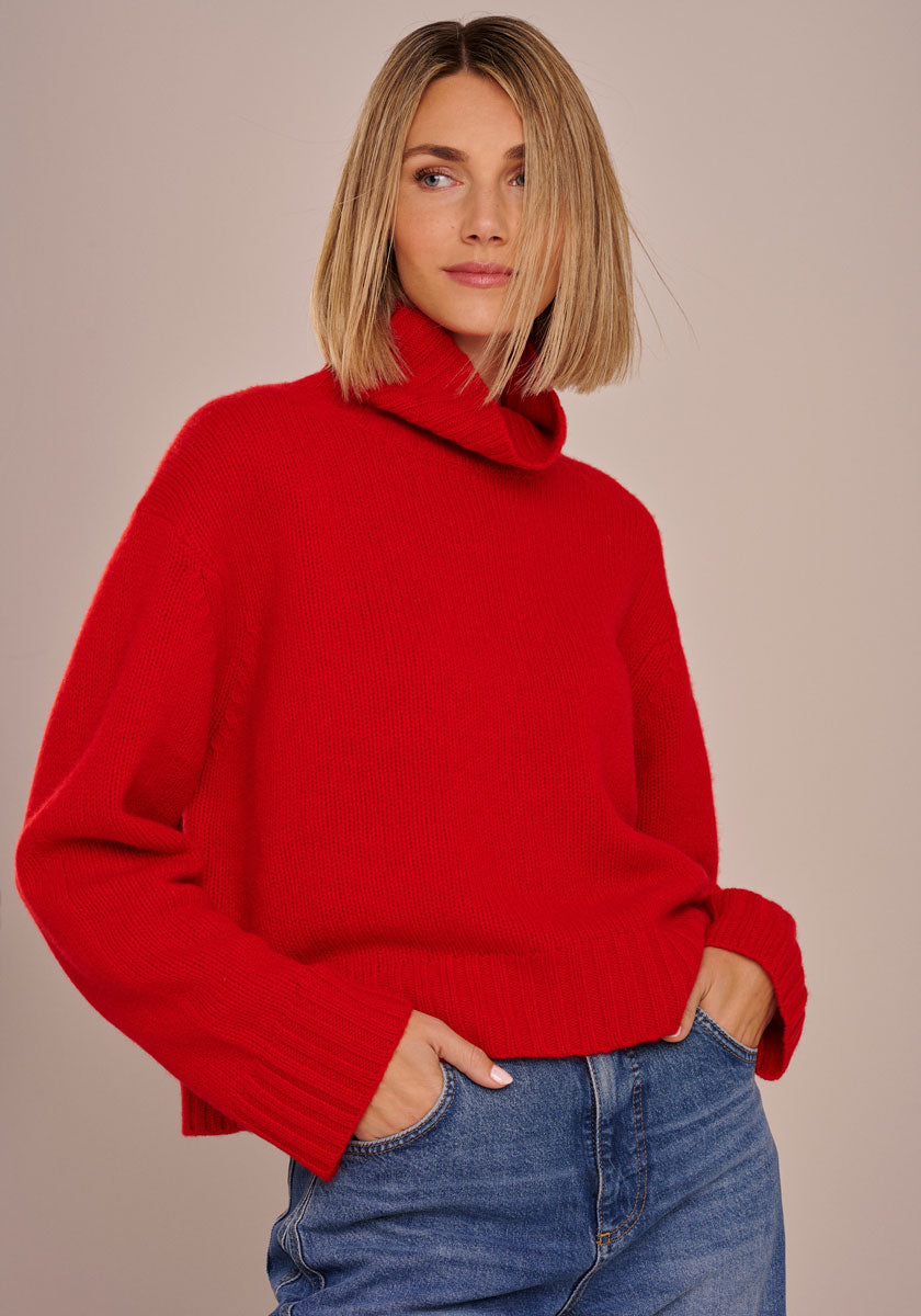 Eine Frau mit glattem, schulterlangem blondem Haar trägt einen locker sitzenden, knallroten Princess Goes Hollywood Rollkragenpullover und blaue Jeans. Sie steht vor einem beigen Hintergrund, die Hände in den Taschen, und schaut ruhig zur Seite.