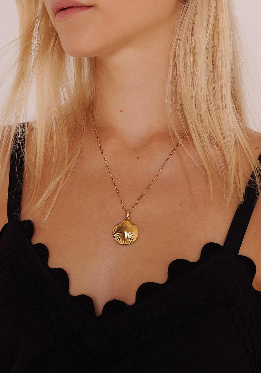 Eine hellhäutige Frau mit hellblondem Haar trägt einen goldenen Princess Goes Hollywood Muschelanhänger an einer feinen Gliederkette. Ihr schwarzes Oberteil ist mit gewellten Wellenkanten versehen, und die sanfte Beleuchtung unterstreicht ihren eleganten Stil.