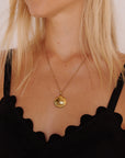 Eine hellhäutige Frau mit hellblondem Haar trägt einen goldenen Princess Goes Hollywood Muschelanhänger an einer feinen Gliederkette. Ihr schwarzes Oberteil ist mit gewellten Wellenkanten versehen, und die sanfte Beleuchtung unterstreicht ihren eleganten Stil.