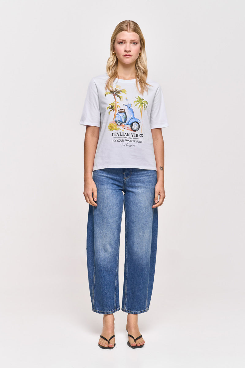 Eine Frau mit langen blonden Haaren blickt nach vorne und trägt ein Princess Goes Hollywood Vespa-Print T-Shirt Baumwolle mit bunten Grafiken und "Italian Vibes", blaue High-Waisted-Weithalsjeans und schwarze Sandalen vor einem schlichten weißen Hintergrund.
