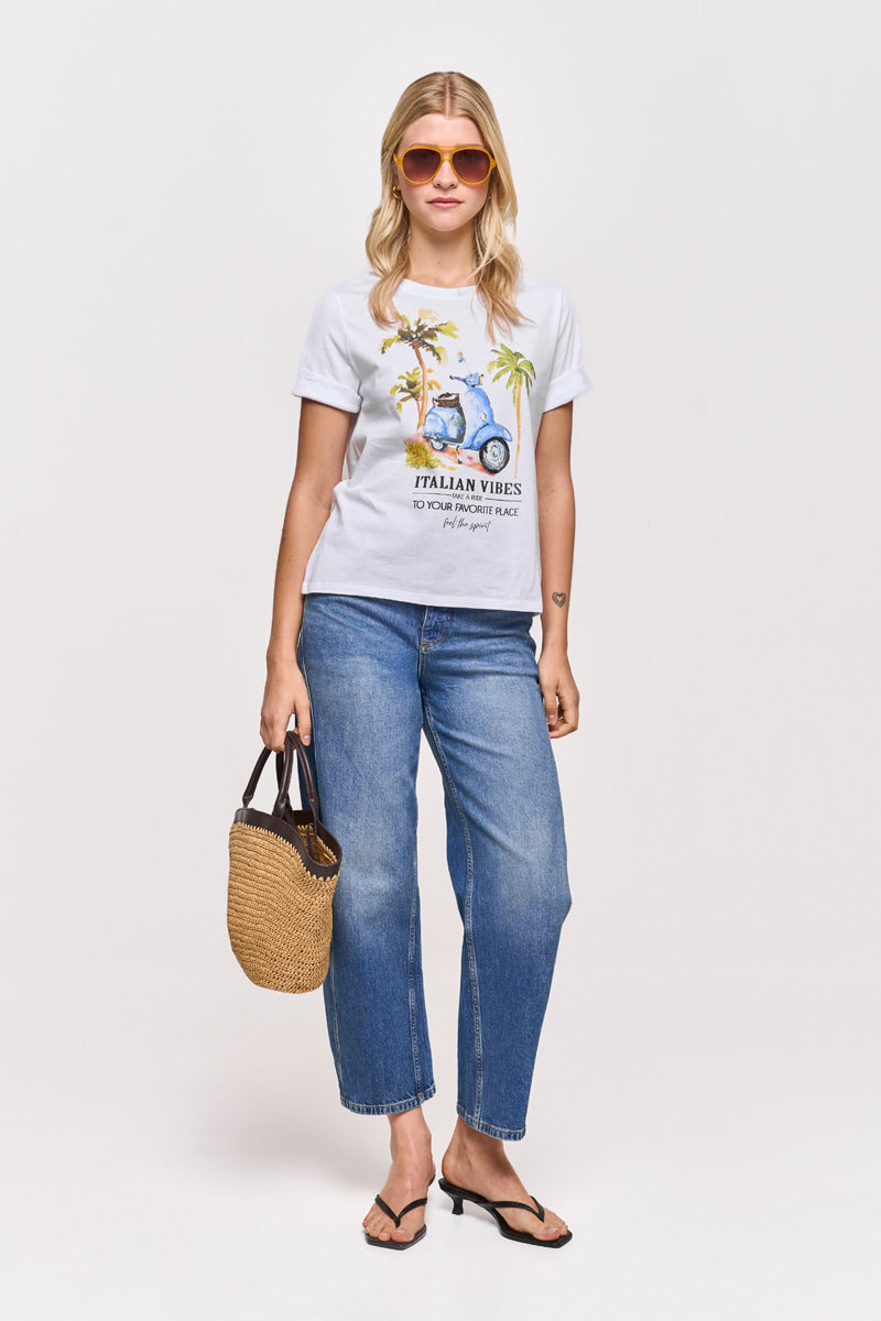 Eine Frau mit blondem Haar trägt eine orangefarbene Sonnenbrille, ein T-Shirt mit dem Aufdruck Princess Goes Hollywood, blaue Jeans, schwarze Flip-Flops und eine geflochtene Strohhandtasche. Sie blickt nach vorne vor einem schlichten weißen Hintergrund.