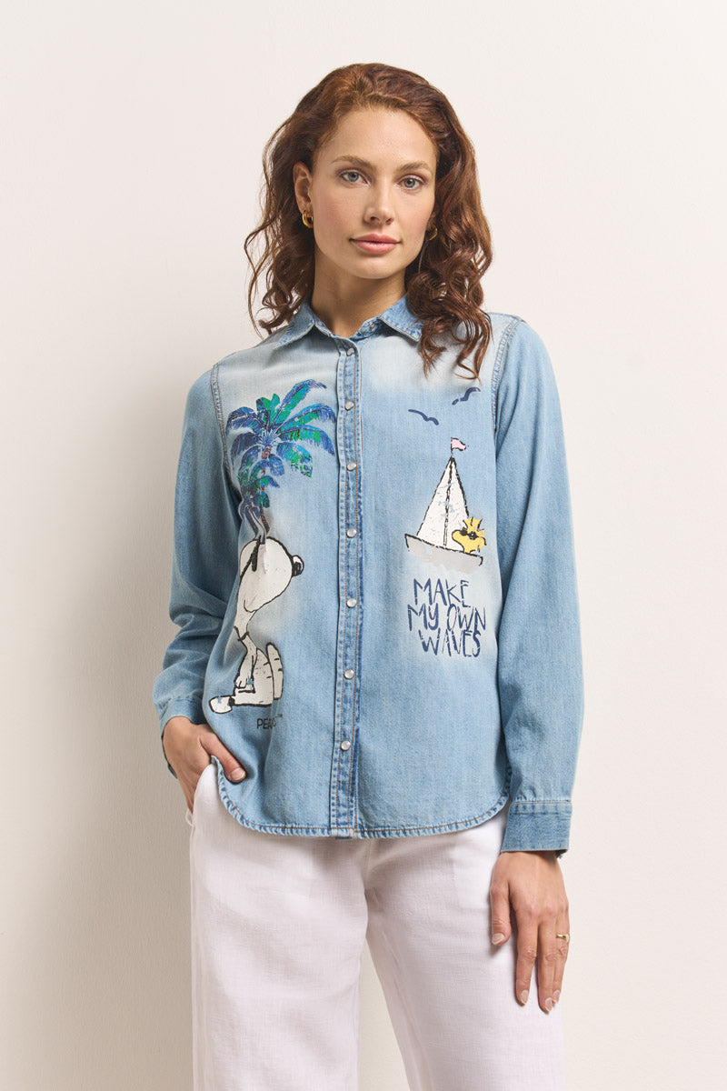 Eine Frau mit gewelltem kastanienbraunem Haar modelliert ein hellblaues Princess Goes Hollywood-Jeanshemd mit Snoopy-Aufdruck, auf dem eine Palme, eine Sonne, ein Segelboot und "MAKE MY OWN WAVES" abgebildet sind, zusammen mit einer weißen Hose vor einer cremefarbenen Wand.