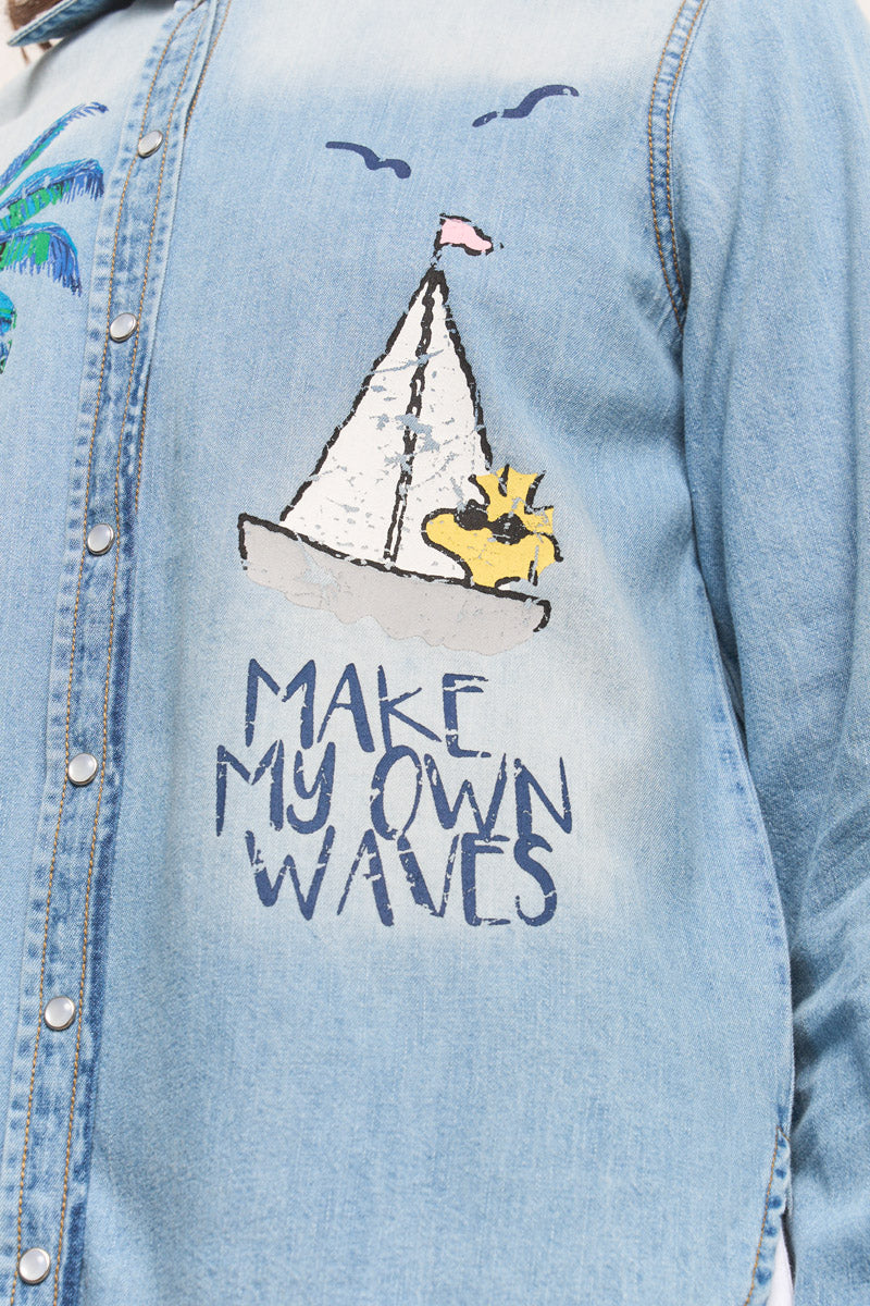 Eine Nahaufnahme eines hellblauen Princess Goes Hollywood Jeans-Shirts aus Baumwolle mit einem verspielten Segelboot, Sonne, Palme und Vögeln als Aufdruck. Darunter steht fett gedruckt "MAKE MY OWN WAVES". Druckknöpfe aus Metall vervollständigen den Look.