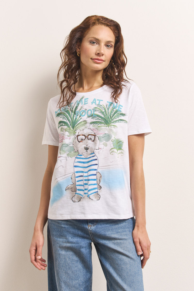 Eine Frau mit gewelltem braunem Haar trägt ein weißes Baumwoll-T-Shirt mit dem Aufdruck "Princess Goes Hollywood", einen glitzernden Hund mit Brille und ein gestreiftes Halstuch, blaue Jeans und steht an einem Pool zwischen Palmen vor einer weißen Wand.