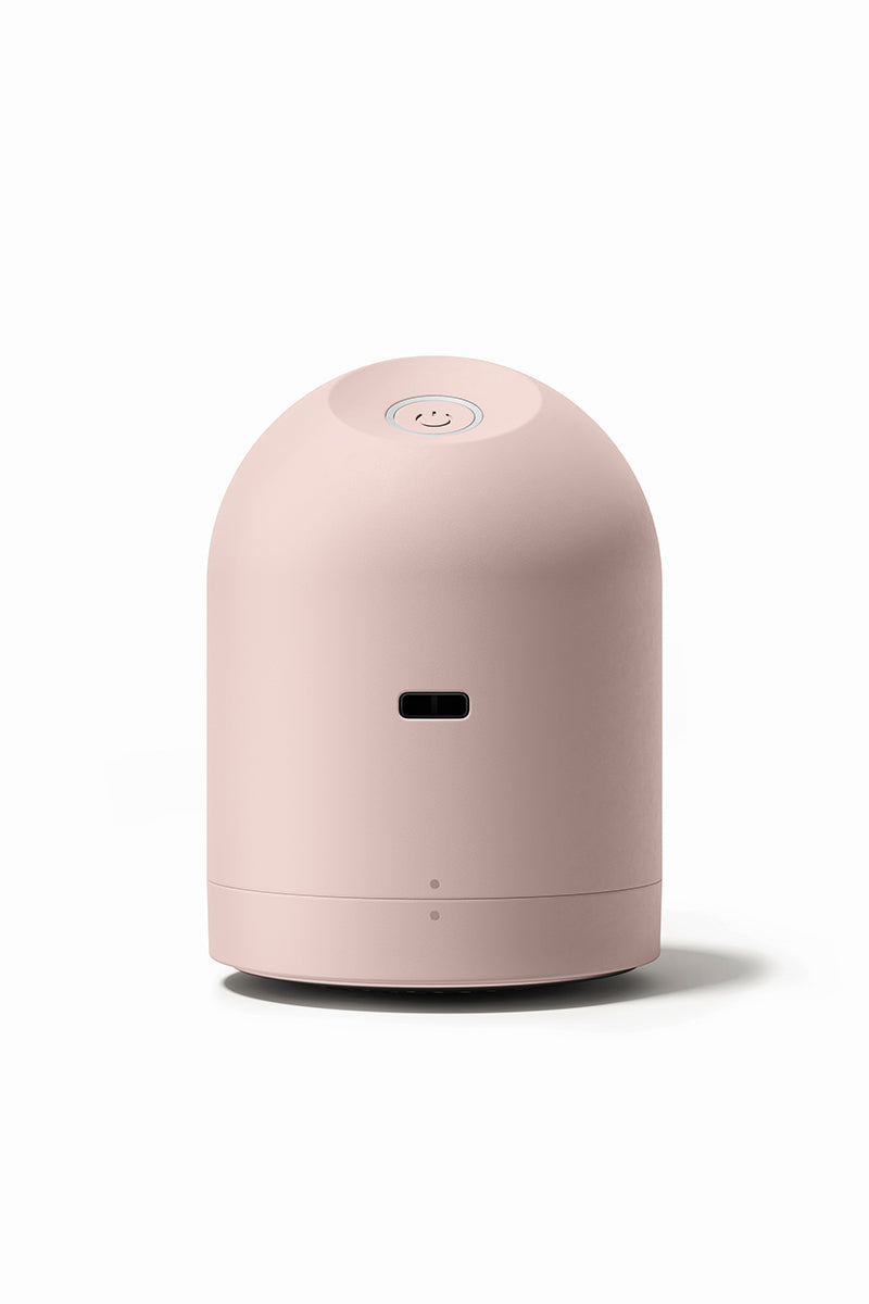 Ein kleiner, kuppelförmiger Steamery Fusselrasierer in mattem Hellrosa mit einer glatten Oberfläche. Er hat einen Smiley-Knopf auf der Oberseite, einen horizontalen Schlitz auf der Vorderseite und eine runde Basis, die alle vor einem weißen Hintergrund abgebildet sind.