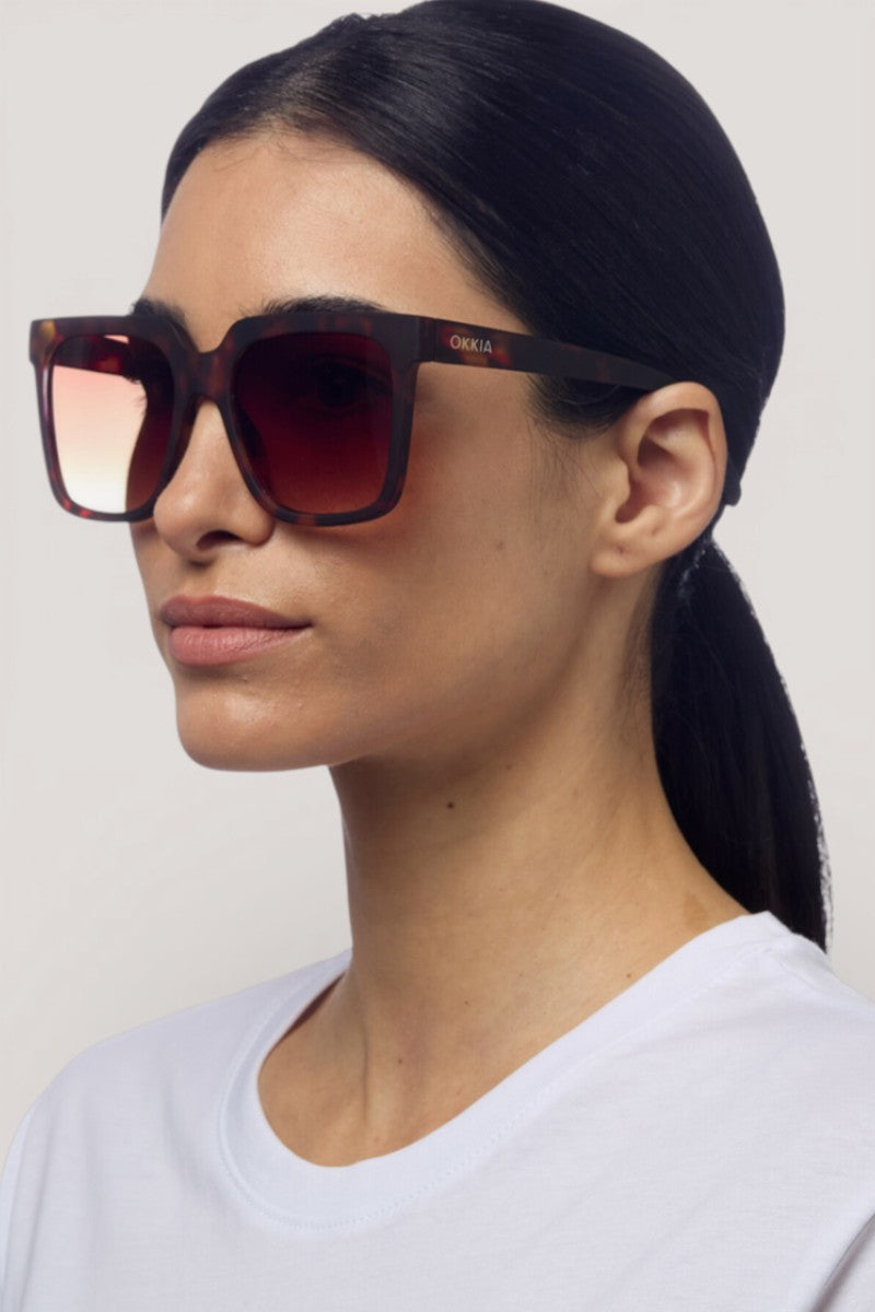 Eine Frau mit glattem schwarzem Haar in einem niedrigen Pferdeschwanz trägt eine übergroße, quadratische Okkia-Sonnenbrille mit braunen Verlaufsgläsern und UV-400-Schutz. Sie blickt leicht zur Seite vor einem schlichten hellgrauen Hintergrund.