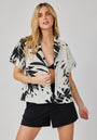 A woman with long blonde hair wears a black crop top, black shorts, and a Bella Dahl airy white short-sleeved button-up shirt with bold black palm print. She stands against a light background, facing forward and holding her shirt slightly open.