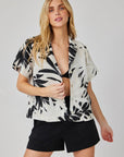 A woman with long blonde hair wears a black crop top, black shorts, and a Bella Dahl airy white short-sleeved button-up shirt with bold black palm print. She stands against a light background, facing forward and holding her shirt slightly open.