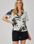 A smiling woman with long blonde hair wears a Bella Dahl short-sleeve, button-up palm print blouse on white fabric with black shorts, standing against a plain light background with one hand on her hip.