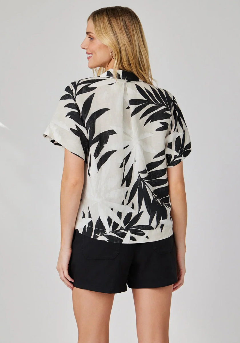 A woman with light hair faces away, wearing a Bella Dahl linen-blend blouse with a bold palm print and black shorts. The simple light gray background accentuates the stylish details of her outfit.