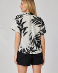A woman with light hair faces away, wearing a Bella Dahl linen-blend blouse with a bold palm print and black shorts. The simple light gray background accentuates the stylish details of her outfit.
