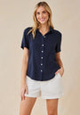 A woman with wavy blonde hair smiles against a neutral background, wearing a navy Bella Dahl Lyocell Kurzarmbluse with Hemdkragen and chest pocket, paired with white high-waisted shorts. She poses casually with her left hand in her pocket.