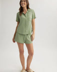 A woman smiles in a light green Bella Dahl Lyocell Kurzarmbluse with Hemdkragen and matching drawstring shorts, paired with beige crisscross slippers. The plain backdrop highlights her relaxed, stylish look.