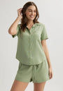 A woman with light skin and long brown hair smiles in a light green Bella Dahl Lyocell short-sleeve blouse with a shirt collar and front pocket, paired with matching drawstring shorts. She stands against a plain light background, hand raised by her head.