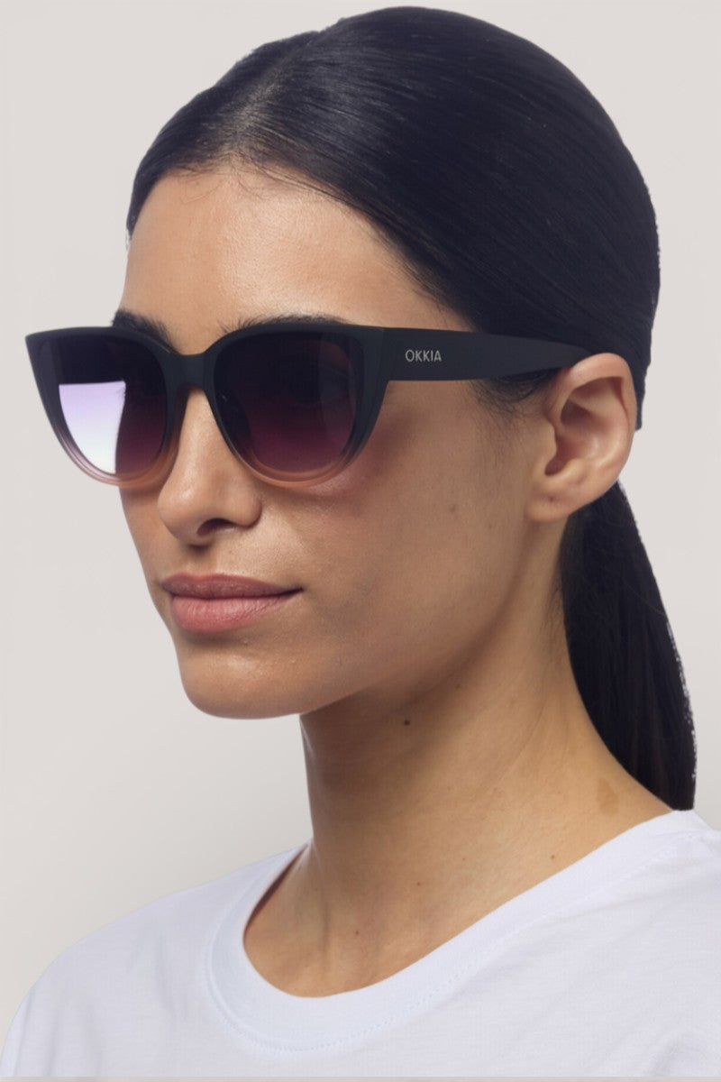 Eine Frau mit glattem, nach hinten gezogenem dunklem Haar trägt eine übergroße schwarze Okkia-Sonnenbrille im schicken Katzenaugen-Design und ein weißes Hemd. Sie blickt nach links, mit ruhigem Ausdruck, vor einem schlichten hellen Hintergrund. Die Verlaufsgläser bieten UV-Schutz.