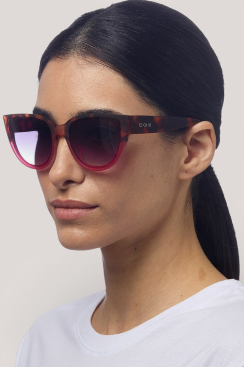 Eine Frau mit glattem, nach hinten gebundenem dunklem Haar trägt eine rote Sonnenbrille mit Farbverlauf und eine schildpattfarbene Katzenaugen-Sonnenbrille von Okkia. Sie hat einen neutralen Gesichtsausdruck, trägt ein schlichtes weißes Hemd und steht vor einem hellgrauen Hintergrund.