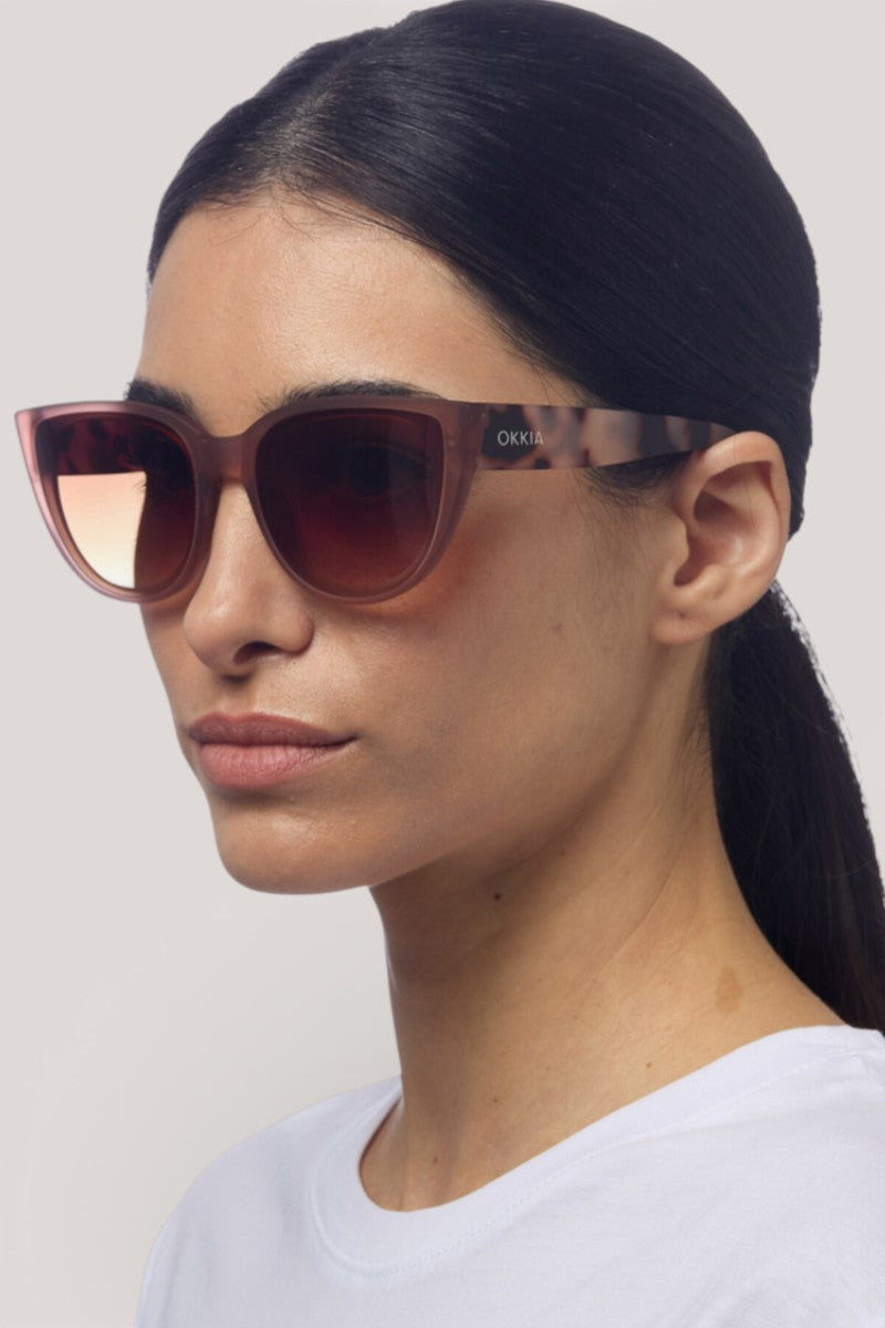 Eine Frau mit glattem, nach hinten gebundenem dunklem Haar trägt eine große Sonnenbrille von Okkia mit leichtem Farbverlauf und gebrandeten Bügeln. Ihre UV-Schutzbrille steht links vor einem schlichten hellen Hintergrund und verleiht ihr ein schickes Flair.