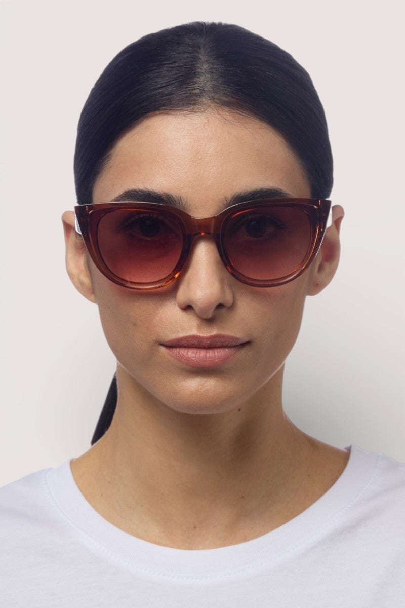 Eine Frau mit glattem, dunklem, zurückgestecktem Haar trägt eine große, braune Okkia-Sonnenbrille und ein weißes T-Shirt mit Rundhalsausschnitt. Sie blickt vor einem schlichten, hellen Hintergrund nach vorne und hat einen neutralen Gesichtsausdruck.