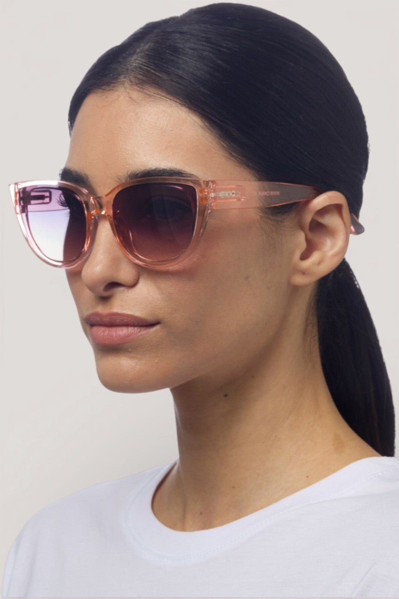 Eine Frau mit glattem, dunklem Haar in einem niedrigen Pferdeschwanz trägt eine rosafarbene, durchsichtige Sonnenbrille mit UV-Schutz von Okkia und ein weißes Hemd. Sie blickt leicht nach rechts vor einem schlichten, hellen Hintergrund, ihr Gesichtsausdruck ist neutral.