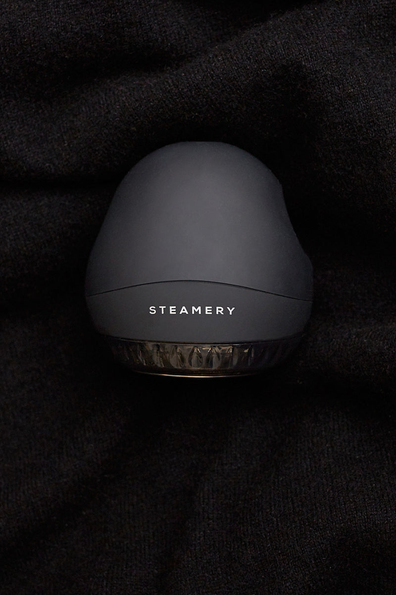 Ein mattschwarzer, ovaler Steamery-Fusselentferner mit dem weißen Schriftzug STEAMERY" sitzt auf einem weichen, dunklen Stoff. Seine glatte Oberseite und der transparente untere Rand unterstreichen das moderne, minimalistische Design des Geräts.