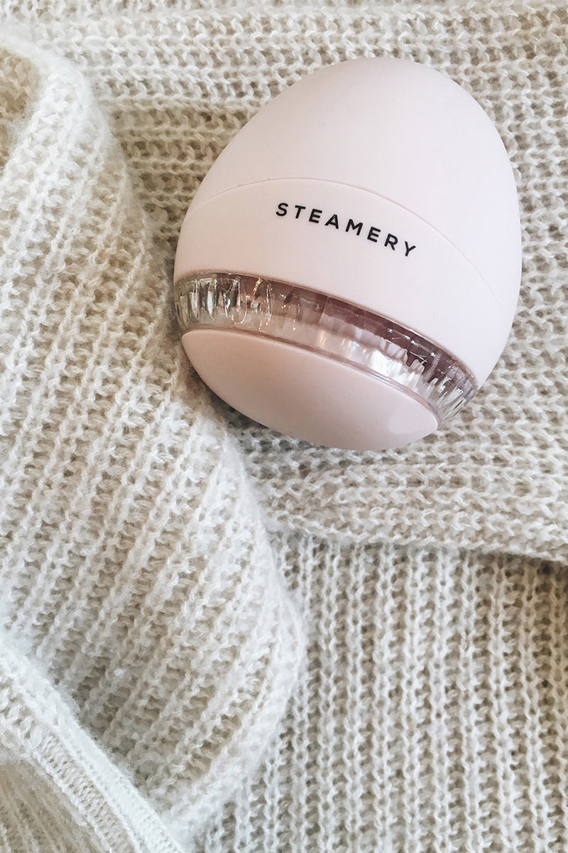 Ein hellrosa, ovaler Steamery Stoffrasierer ruht auf einem cremefarbenen Strickpullover. Der transparente Teil des Pullovers zeigt die Klingen und den aufgesammelten Stoff, während der Rippenstrick für einen gemütlichen Touch sorgt.