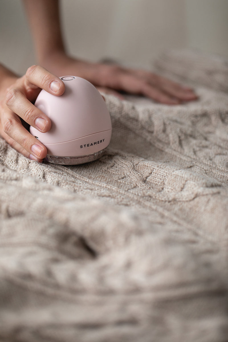 Eine Hand benutzt den rosafarbenen, kuppelförmigen Steamery Fusselrasierer, um Fussel von einem beigen Zopfstrickpullover zu entfernen, wobei ein sanft verschwommener Hintergrund sowohl das Kleidungsstück als auch den eleganten Fusselentferner der Marke Steamery hervorhebt.