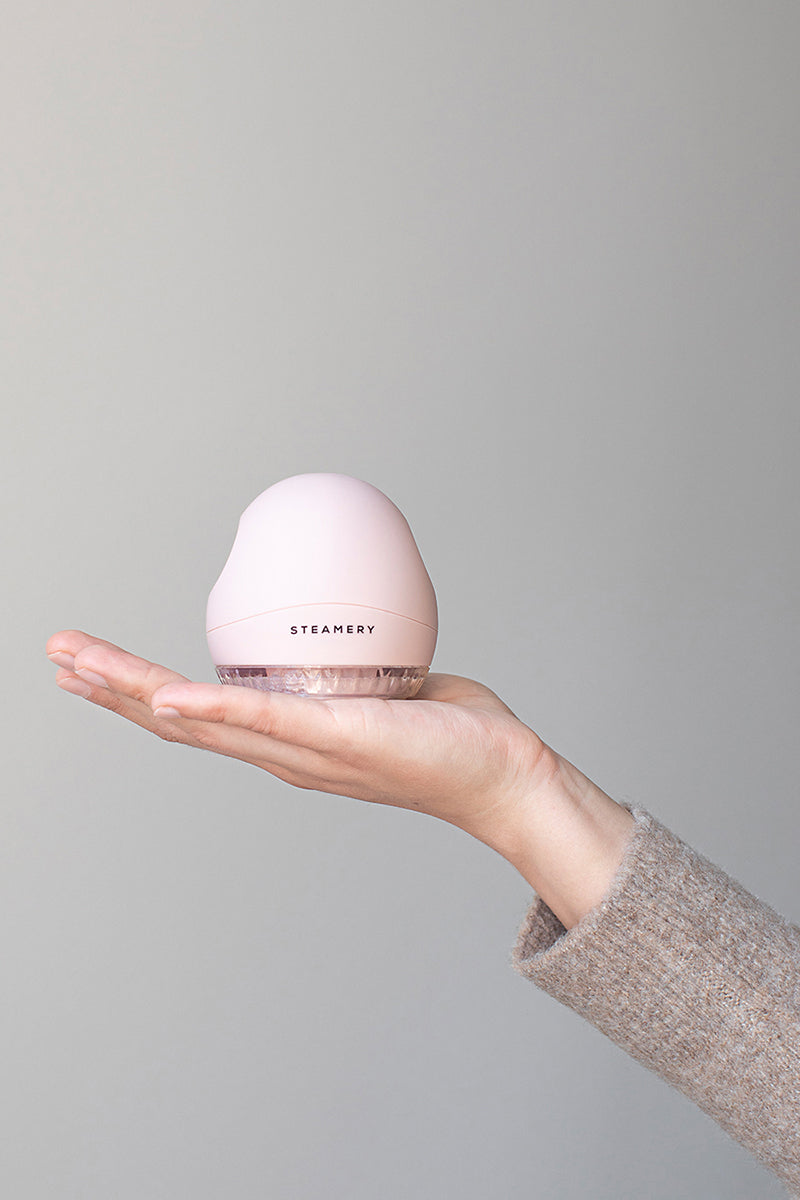 Eine Hand in einem beigen Pullover hält einen blassrosa Steamery Stoffrasierer mit transparentem Boden und abgerundeter Spitze vor einem hellgrauen Hintergrund, der modernes Design und praktische Textilpflege demonstriert.