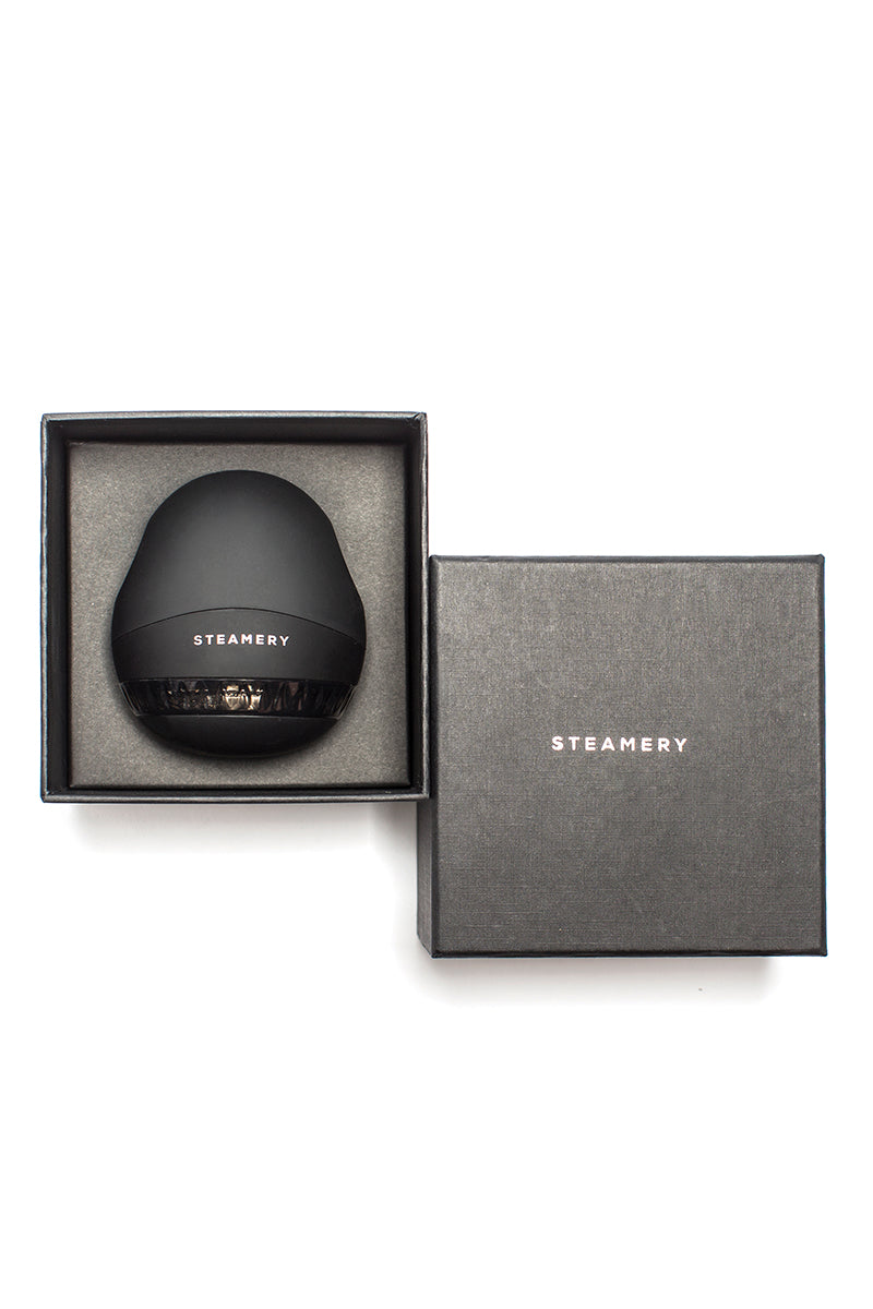 Ein mattschwarzer, eiförmiger Steamery-Rasierer befindet sich in einer offenen schwarzen Schachtel neben dem geschlossenen Deckel; beide tragen den Steamery-Markennamen in weißen Großbuchstaben auf einem sauberen weißen Hintergrund.