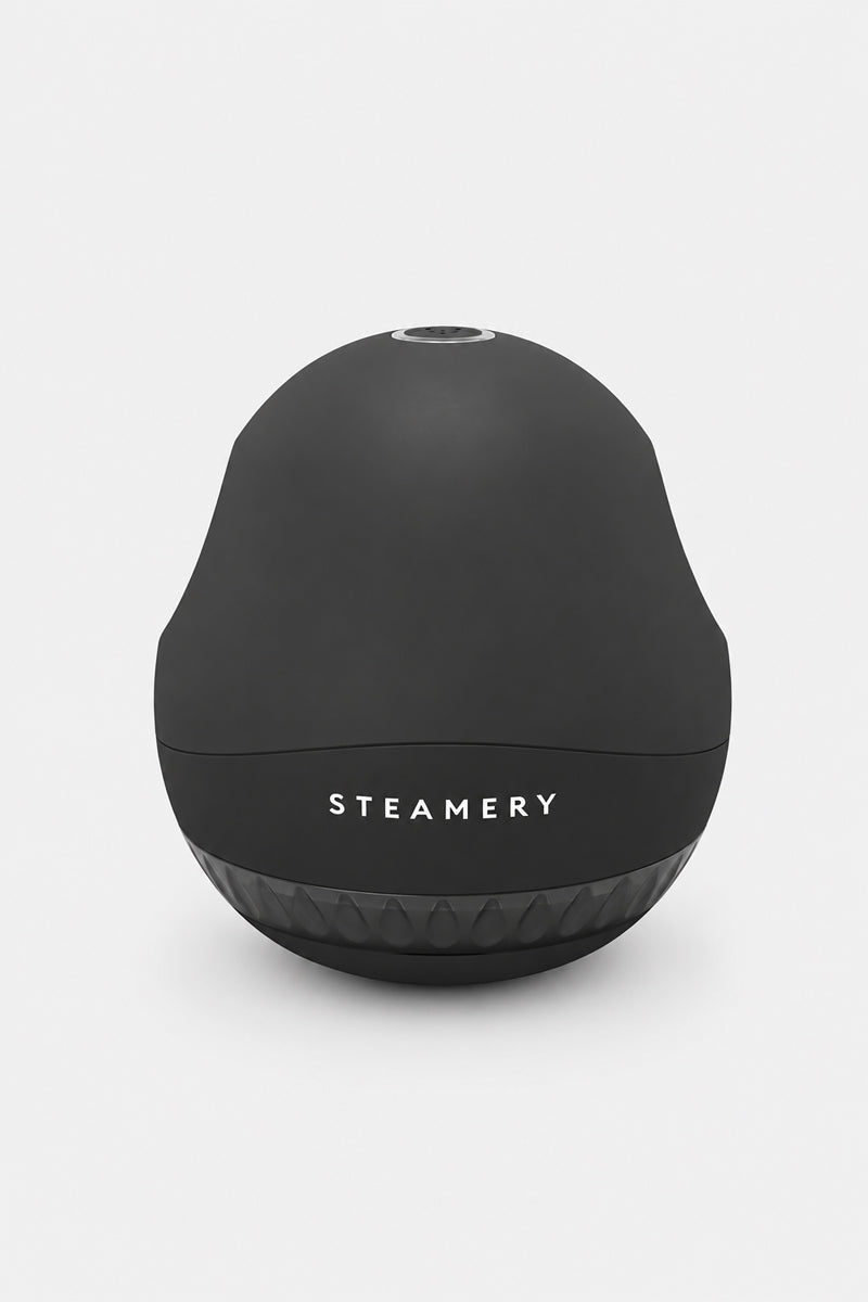 Auf einem weißen Hintergrund ist ein schwarzer, kuppelförmiger Fusselrasierer mit matter Oberfläche zu sehen. Auf der Vorderseite ist das Wort „STEAMERY“ in weißen Großbuchstaben aufgedruckt. An der Unterseite befindet sich ein strukturierter, durchscheinender Bereich zum Auffangen von Stoffflusen, wodurch dieser Stoffrasierer sowohl praktisch als auch stilvoll ist.
